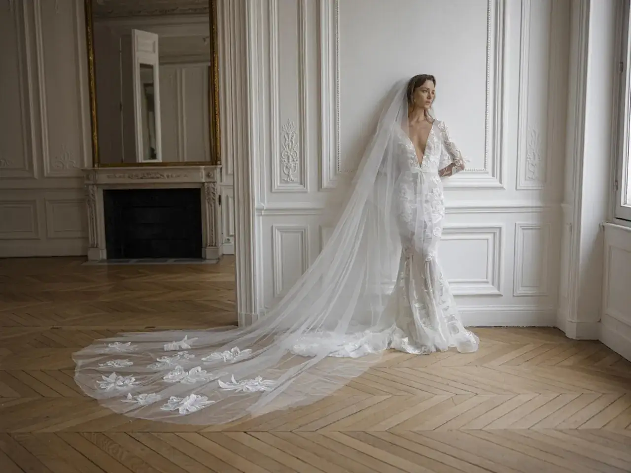 Braut in einem Spitzenkleid mit tiefem Ausschnitt und einem langen, bestickten Schleier, der elegant über den Holzboden fällt. Perfekt für den Schleier Standesamt.