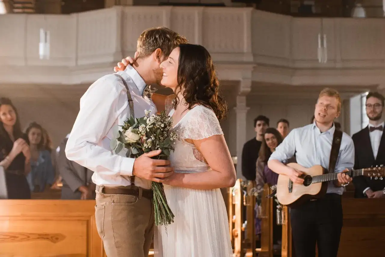 Hochzeitspaar umarmt sich, während ein Mann Gitarre spielt und Gäste zu bekannten Kirchenliedern zum Mitsingen lauschen.