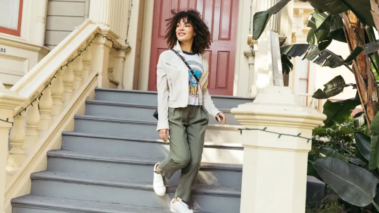 Frau in smart casual Kleidung steigt eine Treppe hinunter. Sie tr&auml;gt eine wei&szlig;e Jacke, ein bedrucktes T-Shirt und gr&uuml;ne Hosen.