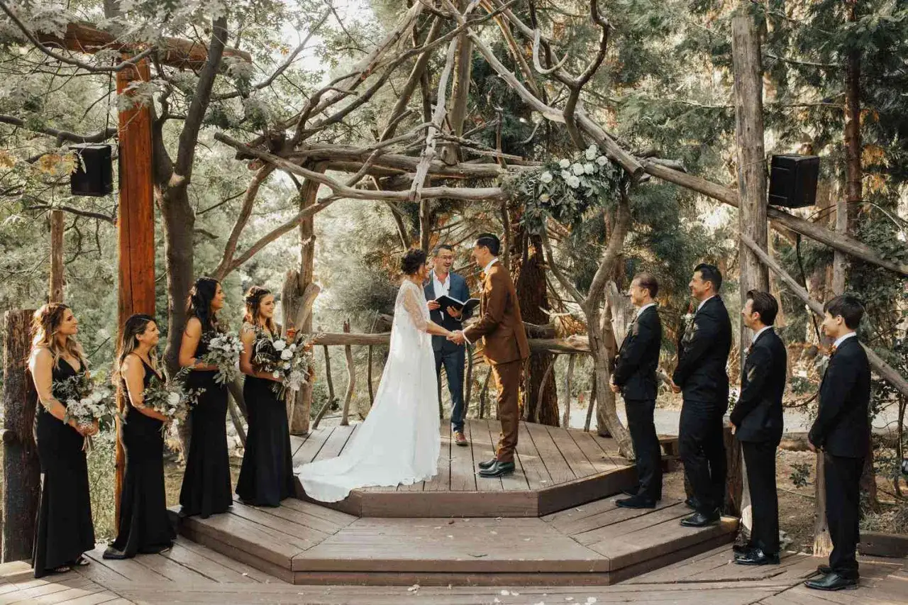 Ein Brautpaar gibt sich das Ja-Wort bei einer romantischen Hochzeit im Wald. Die Zeremonie findet unter einem kunstvollen Holzpavillon statt, umgeben von Bäumen.