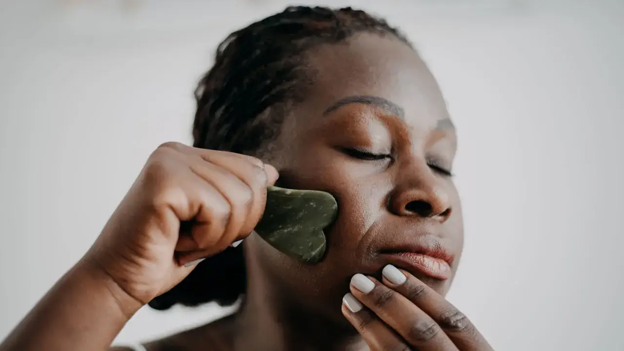 Frau mit geschlossenen Augen nutzt einen grünen Gua Sha Stein zur Gesichtsmassage. Die Gua Sha Stein Wirkung wird hierbei spürbar.