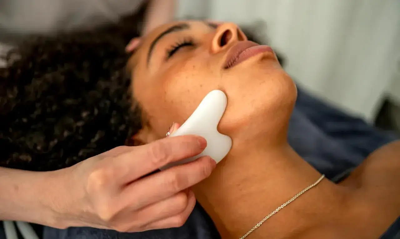 Eine Hand wendet die Gua Sha Technik an einem Gesicht an. Ein weißes Gua Sha Werkzeug gleitet über die Wange einer Frau.