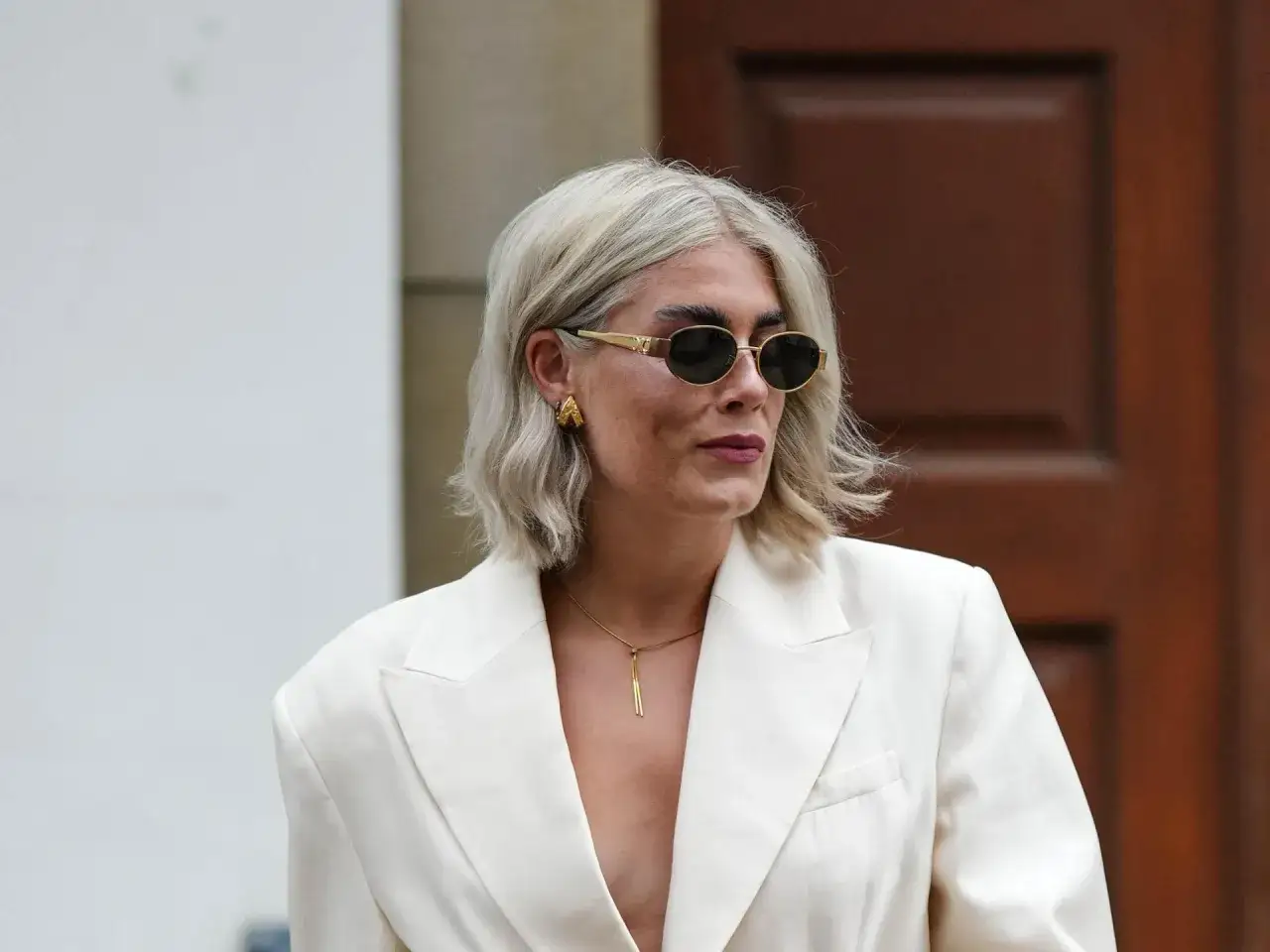 Stilvolle Frau mit shaggy bob ab 50, trägt weißen Blazer, Sonnenbrille und auffälligen Schmuck.