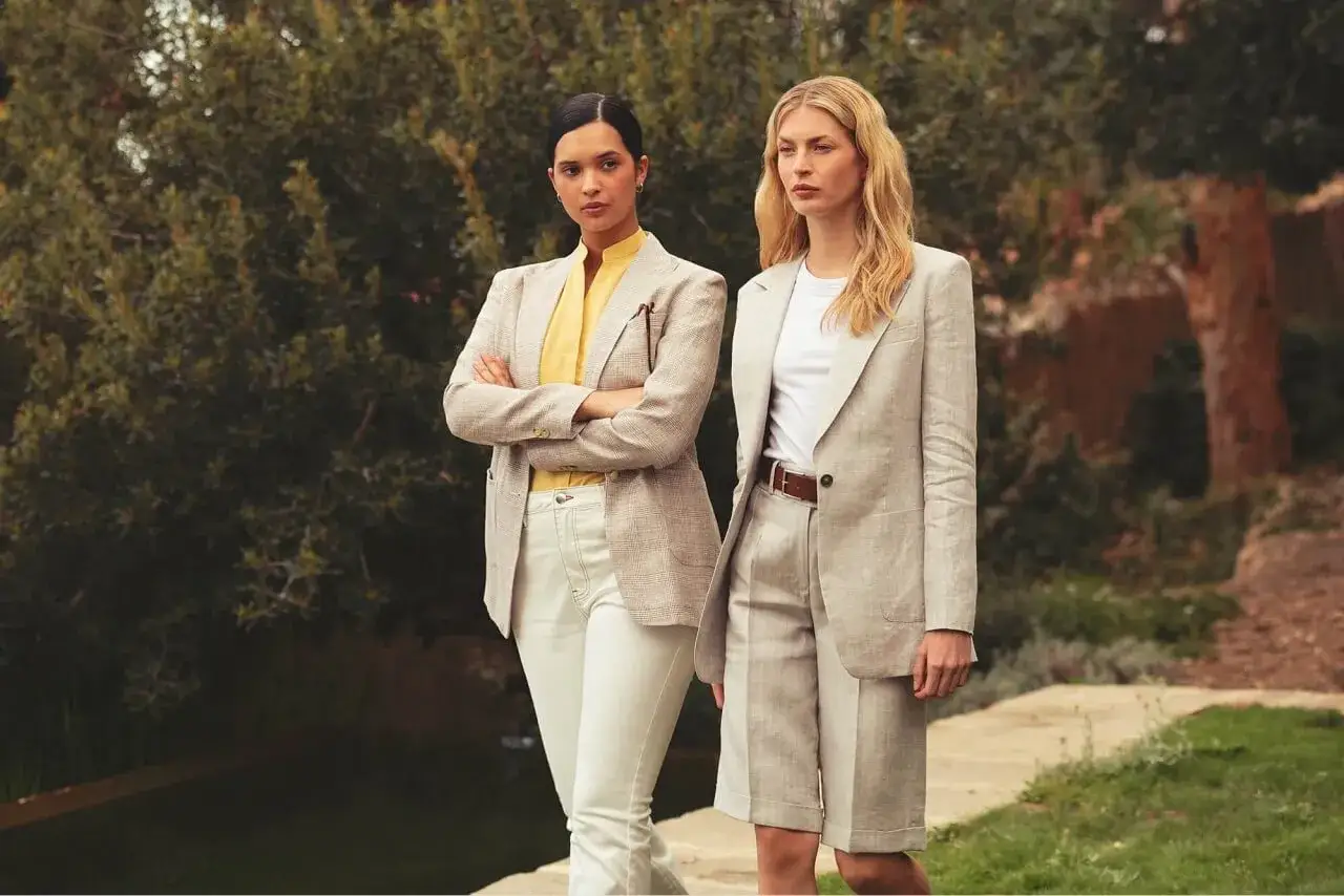 Zwei Frauen in business casual Outfits, eine mit kariertem Blazer und gelbem Hemd, die andere in Leinen-Shorts und Blazer.