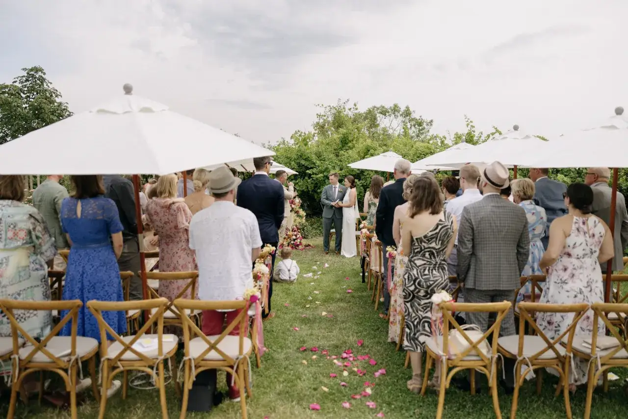 Hochzeit im Weingut: G&auml;ste sitzen auf St&uuml;hlen unter Sonnenschirmen, w&auml;hrend das Brautpaar vor einer &uuml;ppigen Kulisse steht.