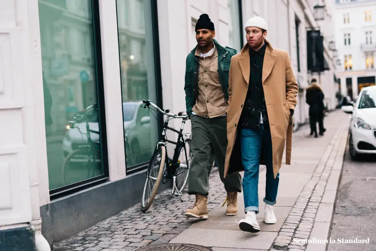 Zwei Männer spazieren mit einem Fahrrad. Sie tragen lässige Outfits, die typisch für skandinavische Modelabels sind: ein Mann im beigen Mantel, der andere in einer grünen Jacke.