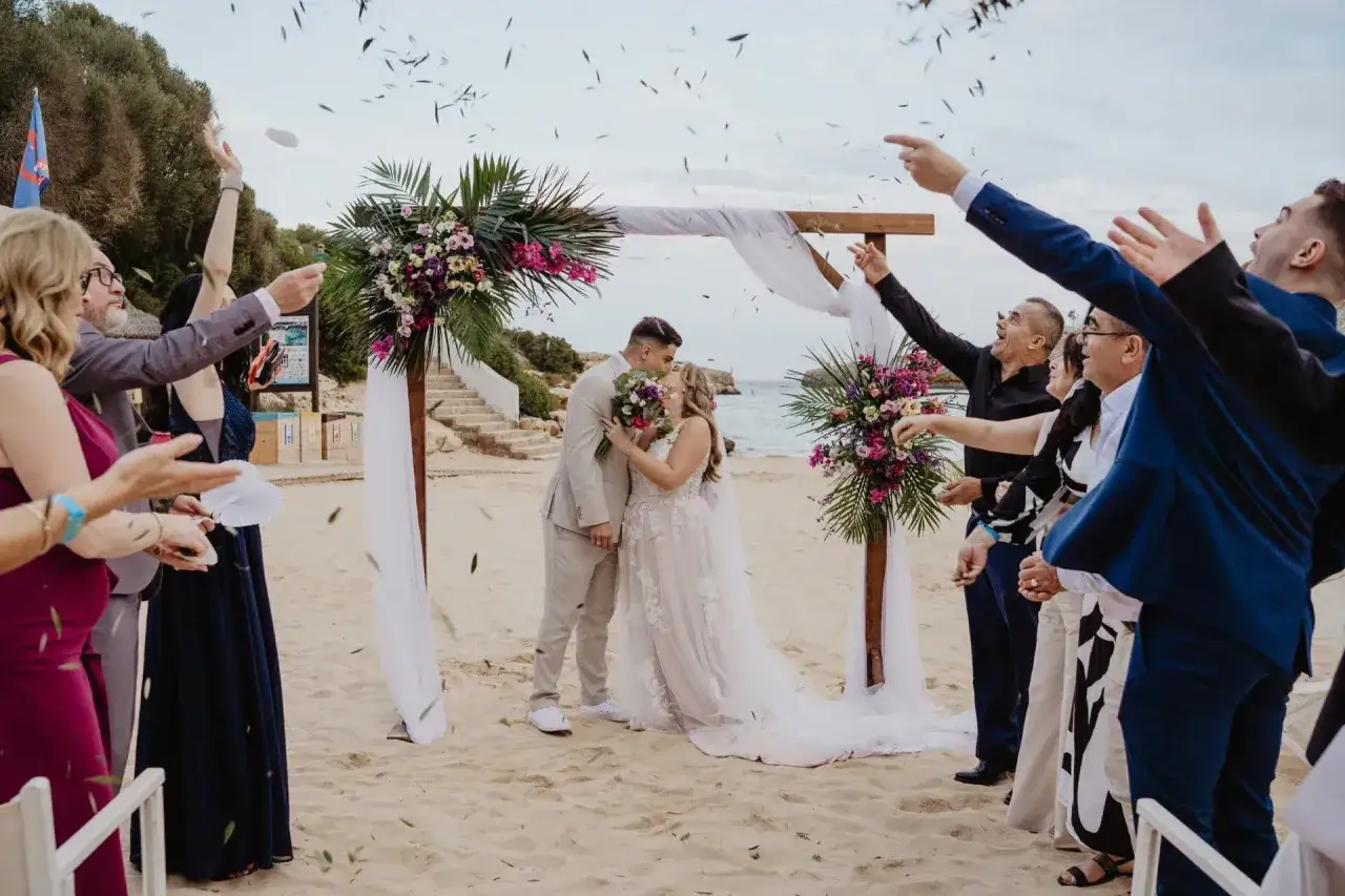Hochzeitspaar küsst sich am Strand, während Gäste Konfetti werfen. Ein traumhaftes Erlebnis, um auf Mallorca am Strand zu heiraten.