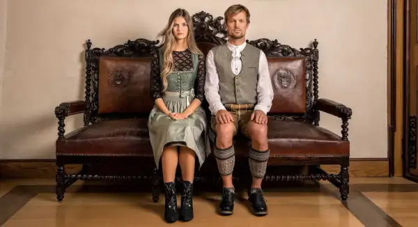 Ein junges Paar sitzt in traditioneller Tracht auf einer antiken Ledercouch. Sie sind bereit, in Tracht zu heiraten und zeigen stolz ihre festliche Kleidung.