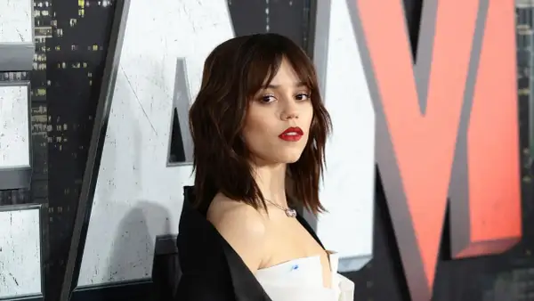 Jenna Ortega mit mittellangem Bob mit Pony und rotem Lippenstift, posiert vor einem großen Logo.