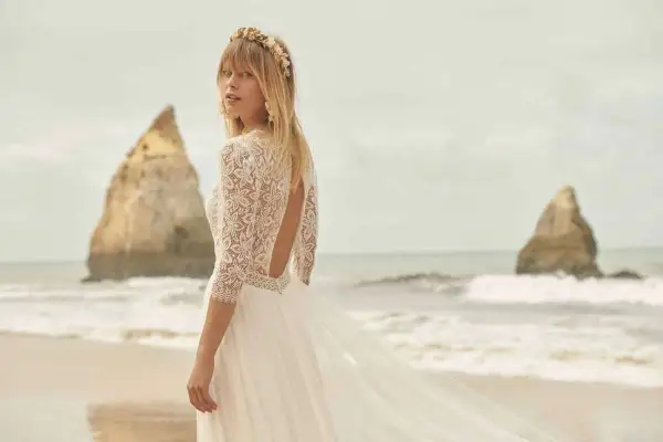 Eine Braut am Strand trägt ein luxuriöses Hochzeitskleid aus Spitze mit offenem Rücken und einem zarten Blumenkranz im Haar.