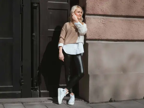 Stylische Frau mit blonden Haaren, die schwarze Lederhose kombiniert mit einem weißen Hemd und einem zweifarbigen Pullover trägt. Weiße Plateau-Sneaker runden den Look ab.