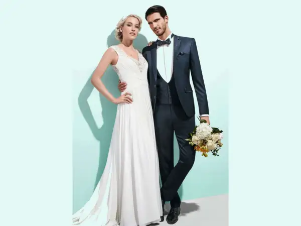Brautpaar posiert für Hochzeitsfoto. Sie trägt ein weißes Kleid, er einen dunklen Anzug mit Weste und Fliege. Der Bräutigam hält einen Blumenstrauß.