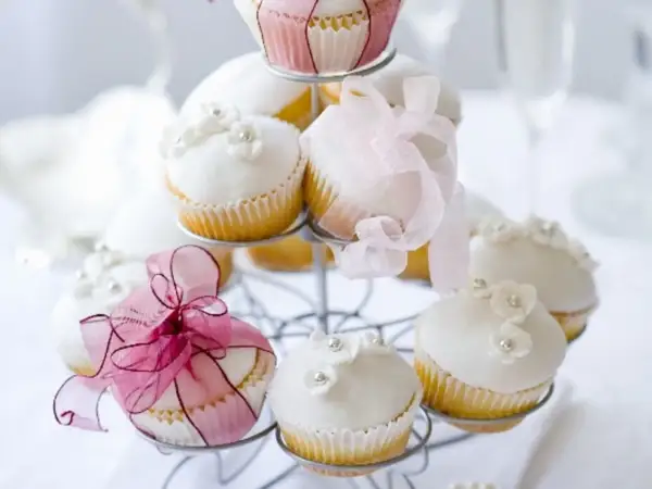 Zarte Muffins mit weißer Glasur, Blüten und Perlen, dekoriert mit rosa Bändern, perfekt für eine Hochzeit.